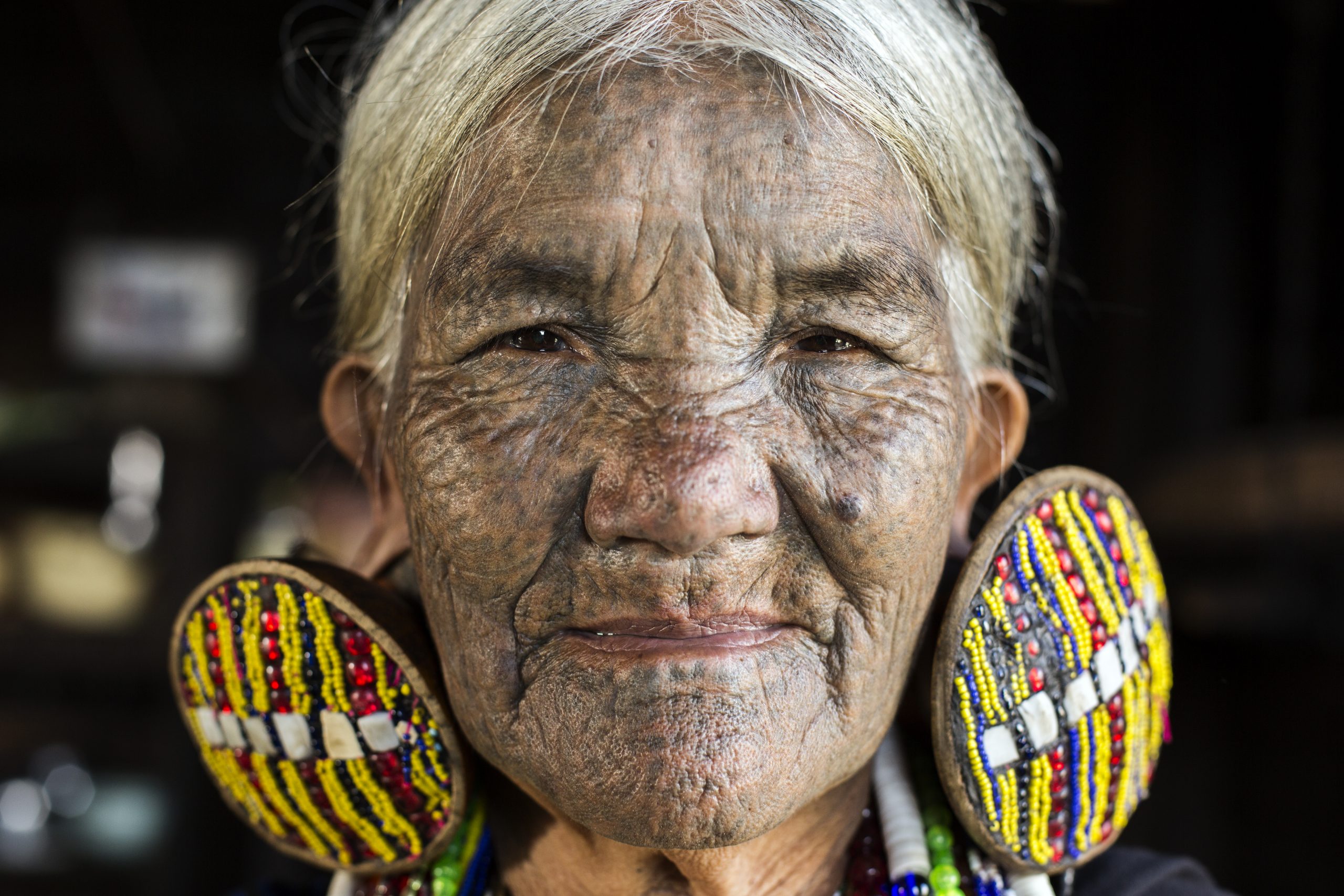 Mindat, Myanmar - December 6th, 2015: Chin tribe tattooed faced woman (Daai) is relaxing at home in Mindat while wearing her exceptionally large traditional earrings and traditional tribal dress. The practice of facial tattooing was outlawed in the 1960's and is usually only seen on old generation women. Chin people, also known as the Kakis are a number of Tibeto-Burman tribal people.