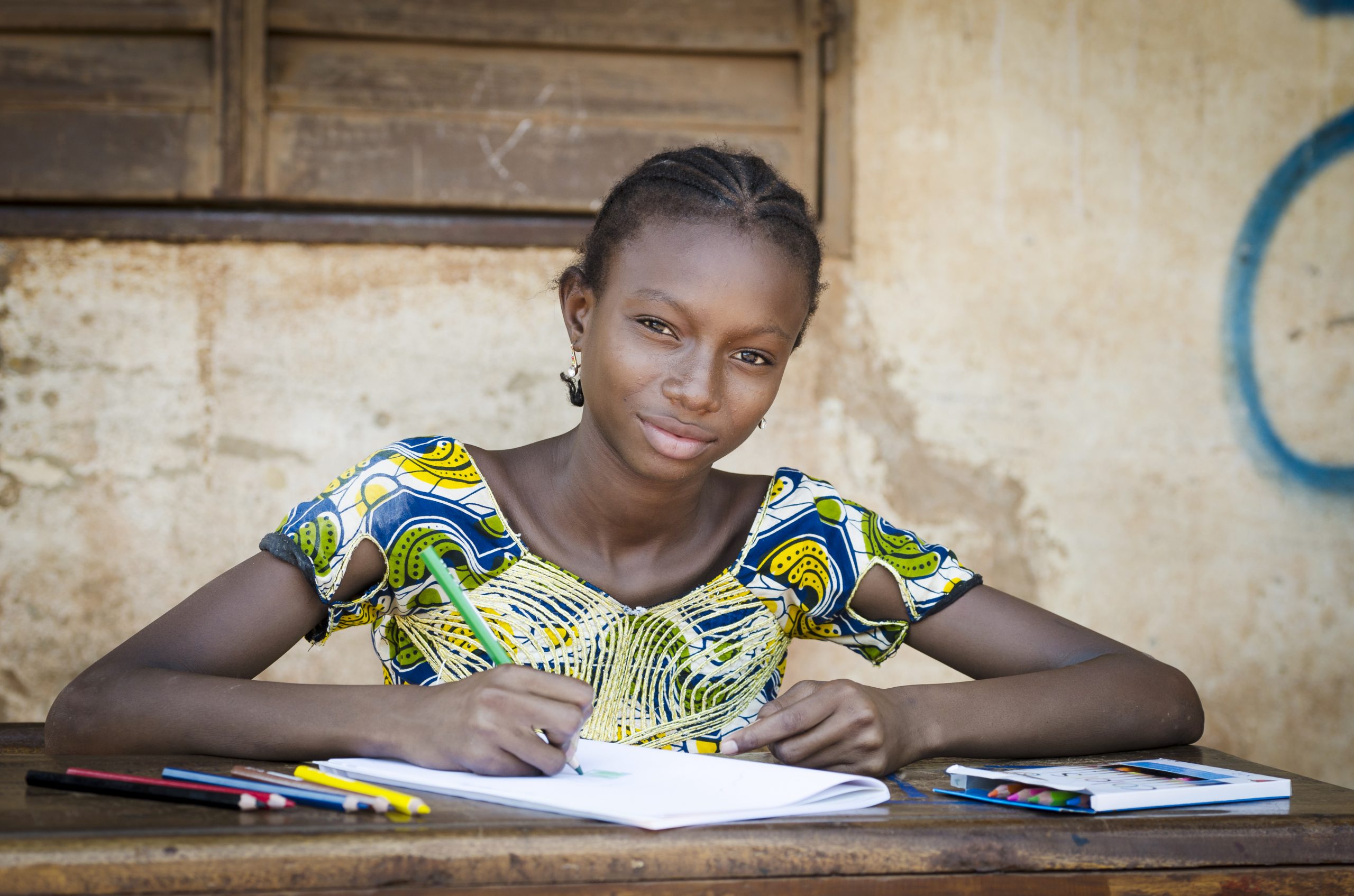 Portrait of an African ethnicity schoolgirl (age 13) in an educational environment in Bamako, Mali learning her lesson outdoors sitting on a desk and slightly smiling.