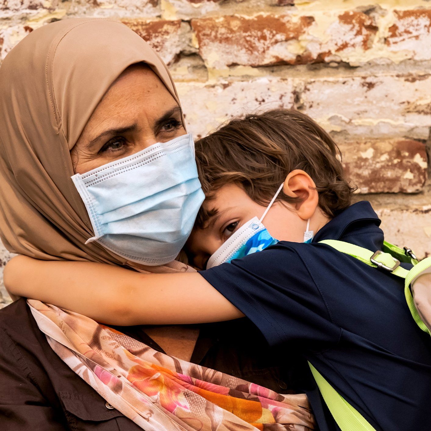 Mature caucasian muslim woman wearing hijab, both wearing protective face masks, carrying her son back to school, on a brick wall background