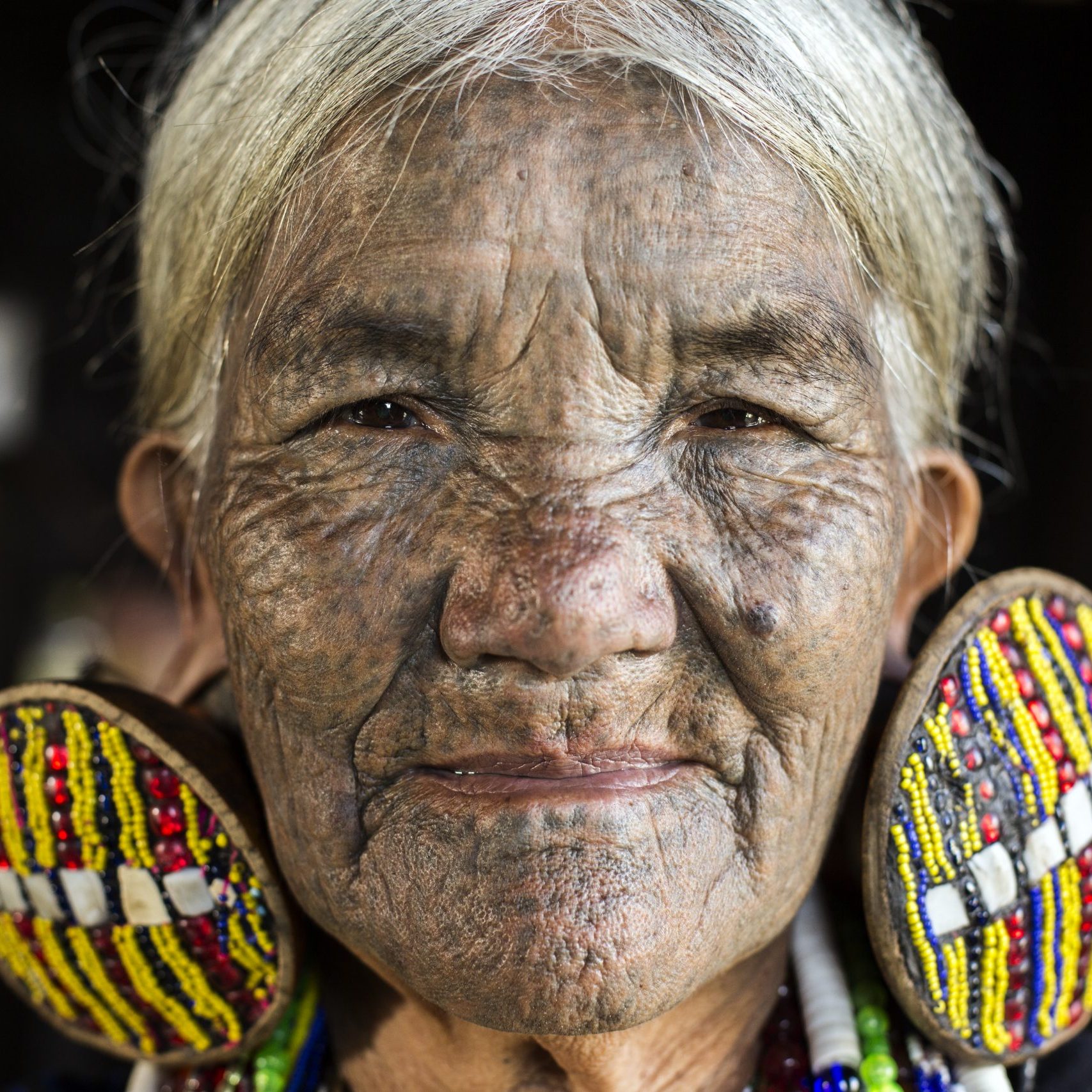 Mindat, Myanmar - December 6th, 2015: Chin tribe tattooed faced woman (Daai) is relaxing at home in Mindat while wearing her exceptionally large traditional earrings and traditional tribal dress. The practice of facial tattooing was outlawed in the 1960's and is usually only seen on old generation women. Chin people, also known as the Kakis are a number of Tibeto-Burman tribal people.
